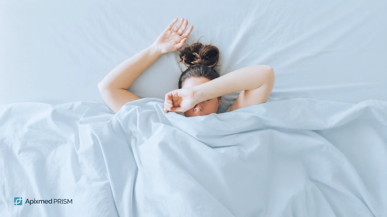 Impact of Biorhythm Disruptions on Sleep Quality and Fatigue — Apixmed Prism A woman in bed covering her face with her arm under the blanket, illustrating chronic fatigue, difficult morning waking, and disrupted circadian patterns.